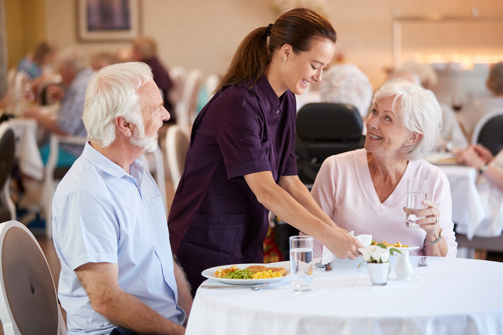 A cheerful staff member serves up dinner to two senior residents